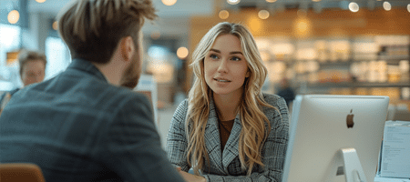 Dos profesionales del marketing en una conversación estratégica en un entorno de oficina moderna. Están sentados frente a un monitor de computadora, enfocados en discutir detalles importantes de su proyecto. La mujer, con una expresión atenta y segura, escucha al hombre mientras intercambian ideas. La oficina está bien iluminada, con un diseño contemporáneo que incluye estanterías y una atmósfera acogedora. Esta interacción refleja un ambiente de trabajo colaborativo y orientado a objetivos, donde la comunicación y la estrategia son clave para el éxito del equipo.